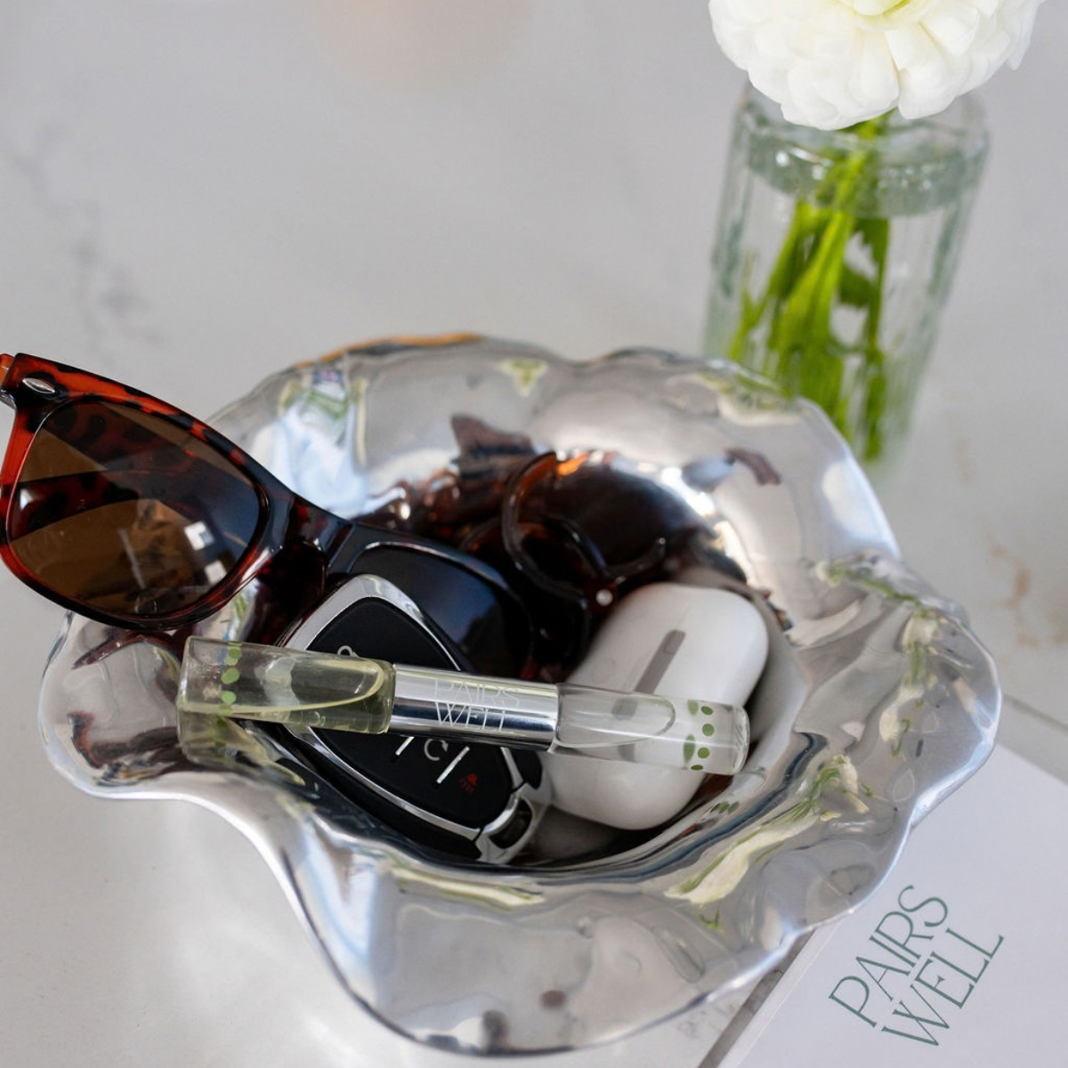 A scalloped clear acrylic or glass tray holding sunglasses, a car key fob, white earbud case, and a small perfume sample vial, styled on a light marble-like surface with a white flower in a glass vase in the background.