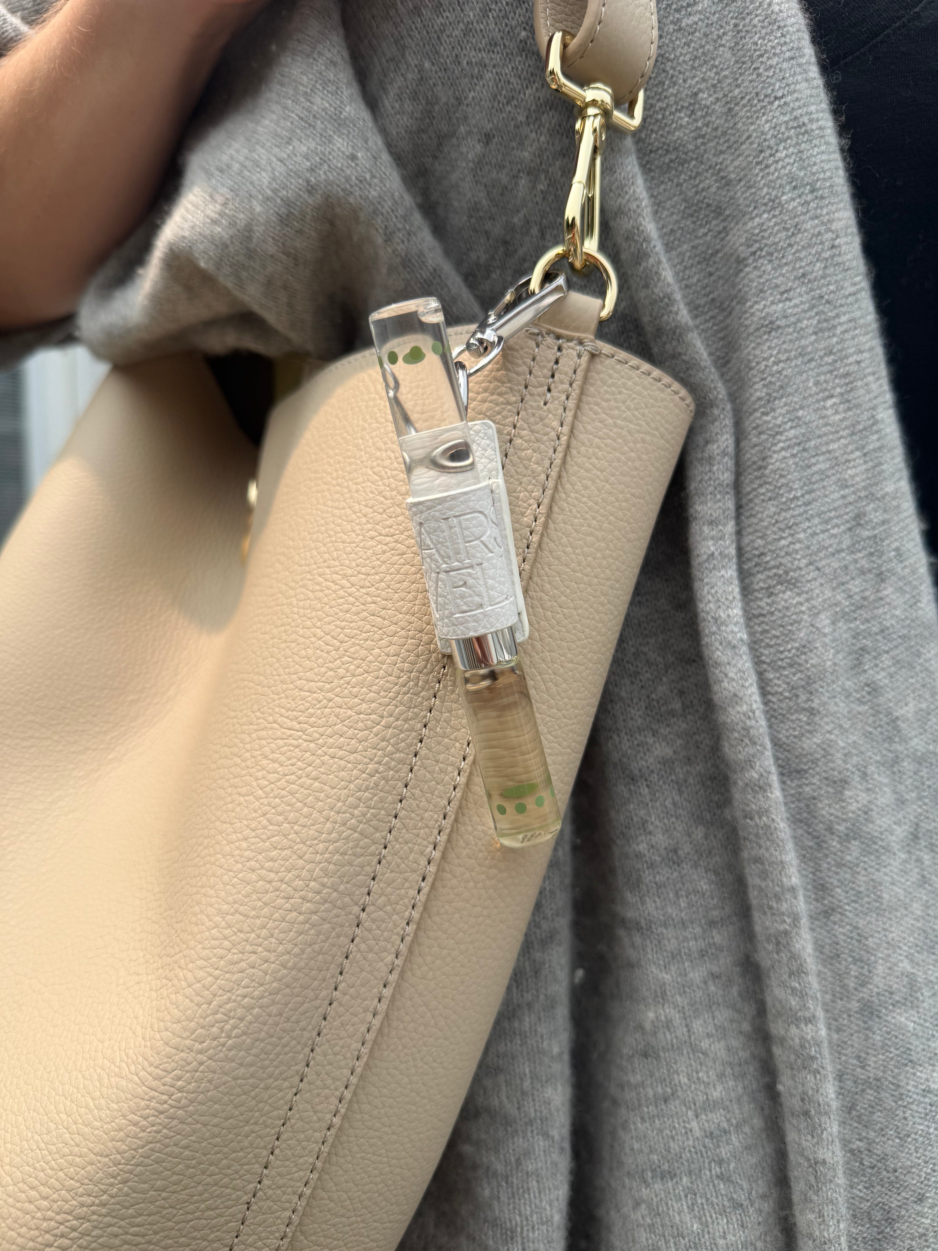 Close-up of a beige pebbled leather shoulder bag with a transparent plastic clip and gold-tone hardware, worn over a gray knit sweater.